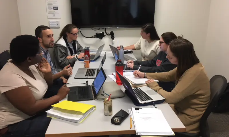 Six students at a table in a conference room working on a project together on their laptops